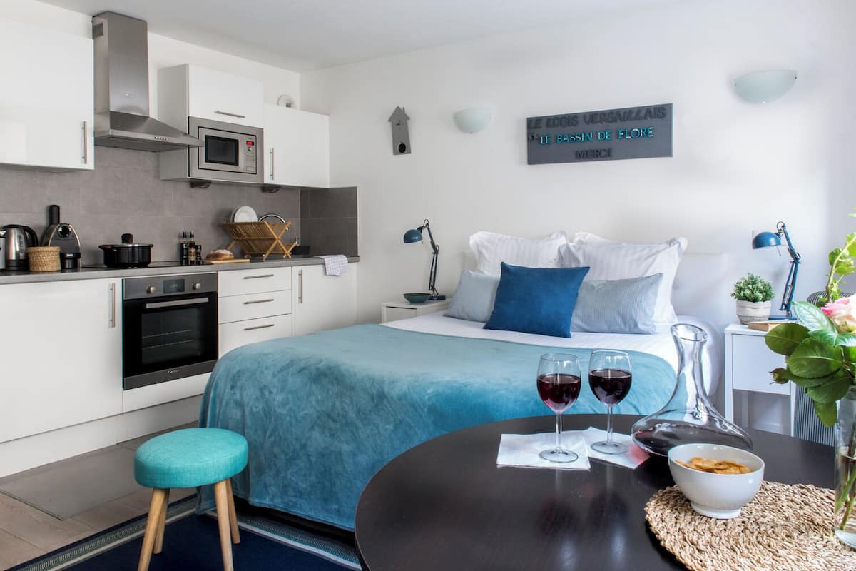 Una habitación de hotel moderna con kitchenette, una cama con ropa de cama azul, una mesita de noche con copas de vino y un florero con flores.
