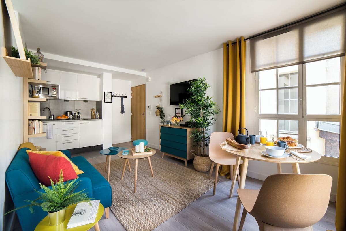 Un espacio habitable compacto con kitchenette, un comedor con productos para el desayuno, un sofá y un gran ventanal con cortinas.
