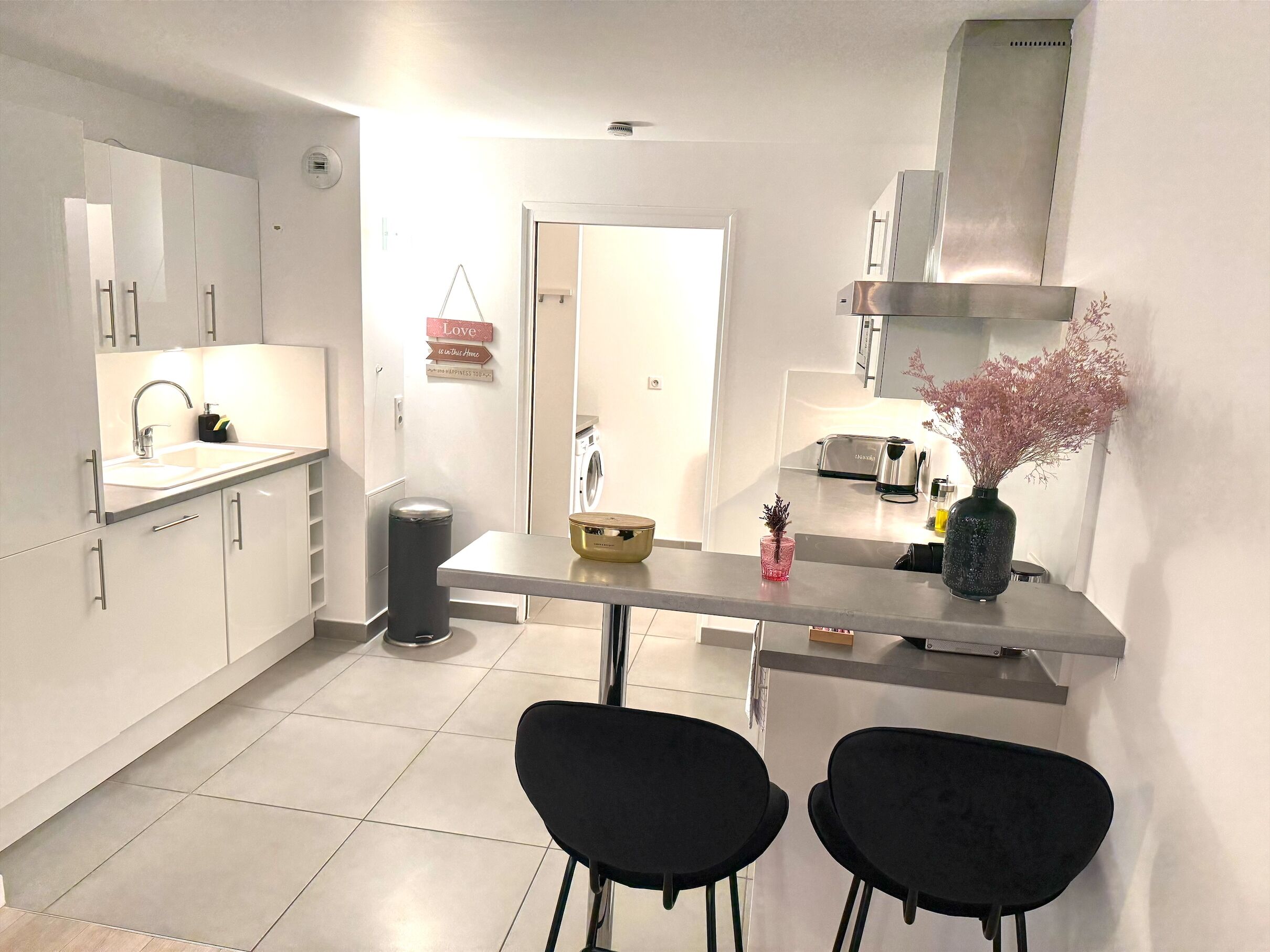 Una cocina moderna con armarios blancos, una campana extractora de acero inoxidable, un lavamanos y un comedor con dos sillas negras.