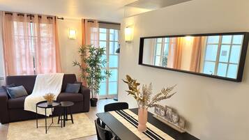 A modern living room with a sofa, coffee table, and dining table. There are potted plants, a mirror, and wall-mounted lights.