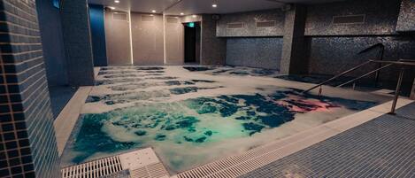 Una piscina interior moderna con superficie de agua cristalina, paredes de azulejos y una escalera metálica que baja.