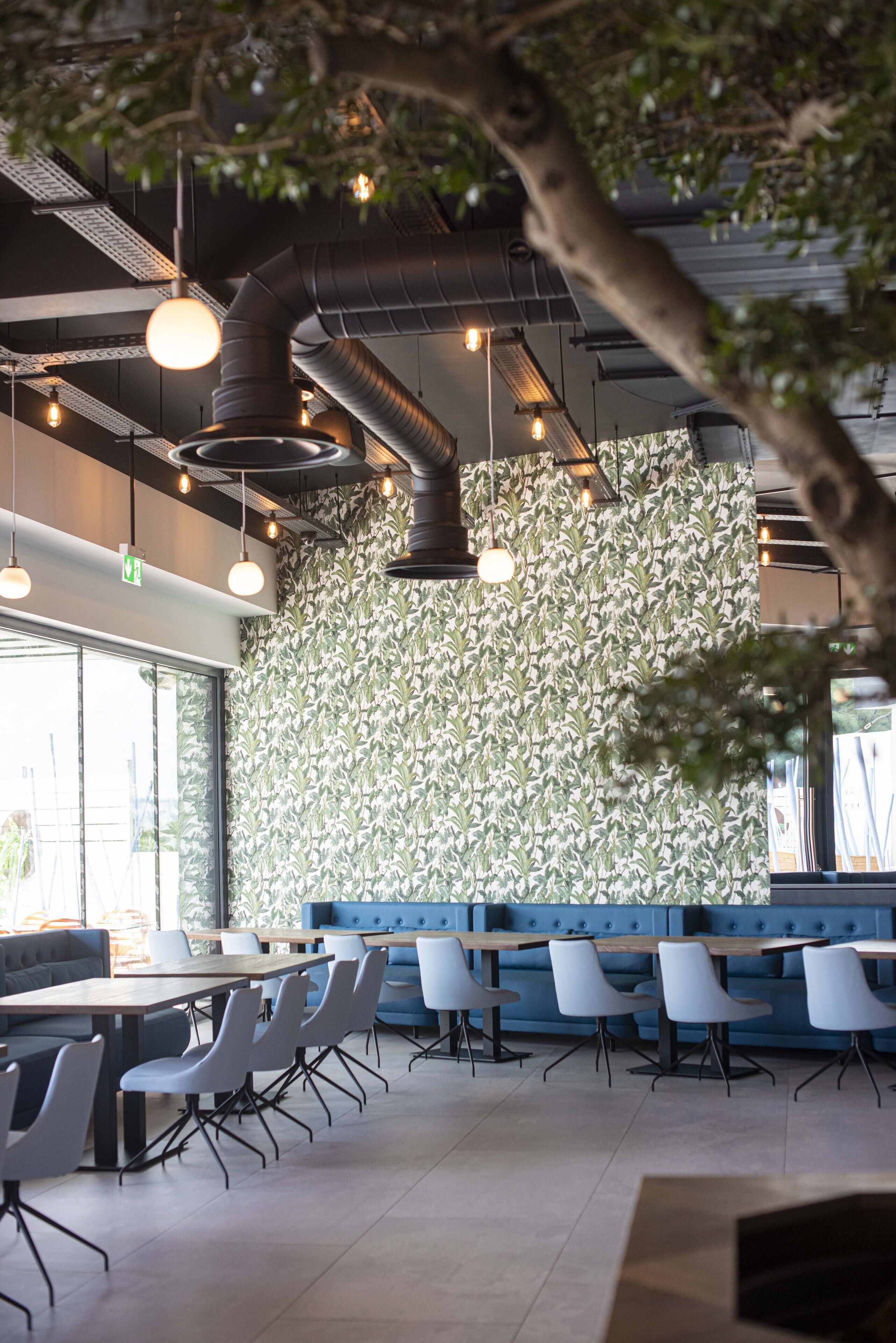 Un espace repas moderne avec un grand mur recouvert d’un papier peint à motifs, un coin salon avec des banquettes bleues et une longue table entourée de chaises.