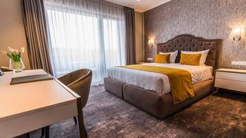 A hotel room with a large bed, a desk, and a chair. There is a window with curtains and a wall-mounted lamp.