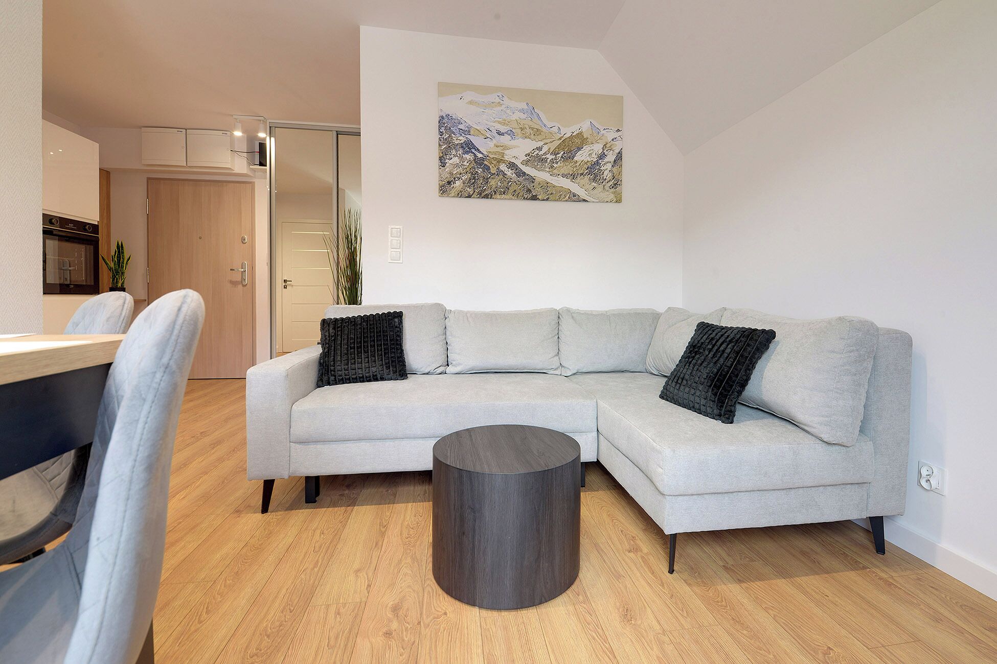 A modern living room with a grey sectional sofa, a round coffee table, and a painting of a mountain landscape on the wall.
