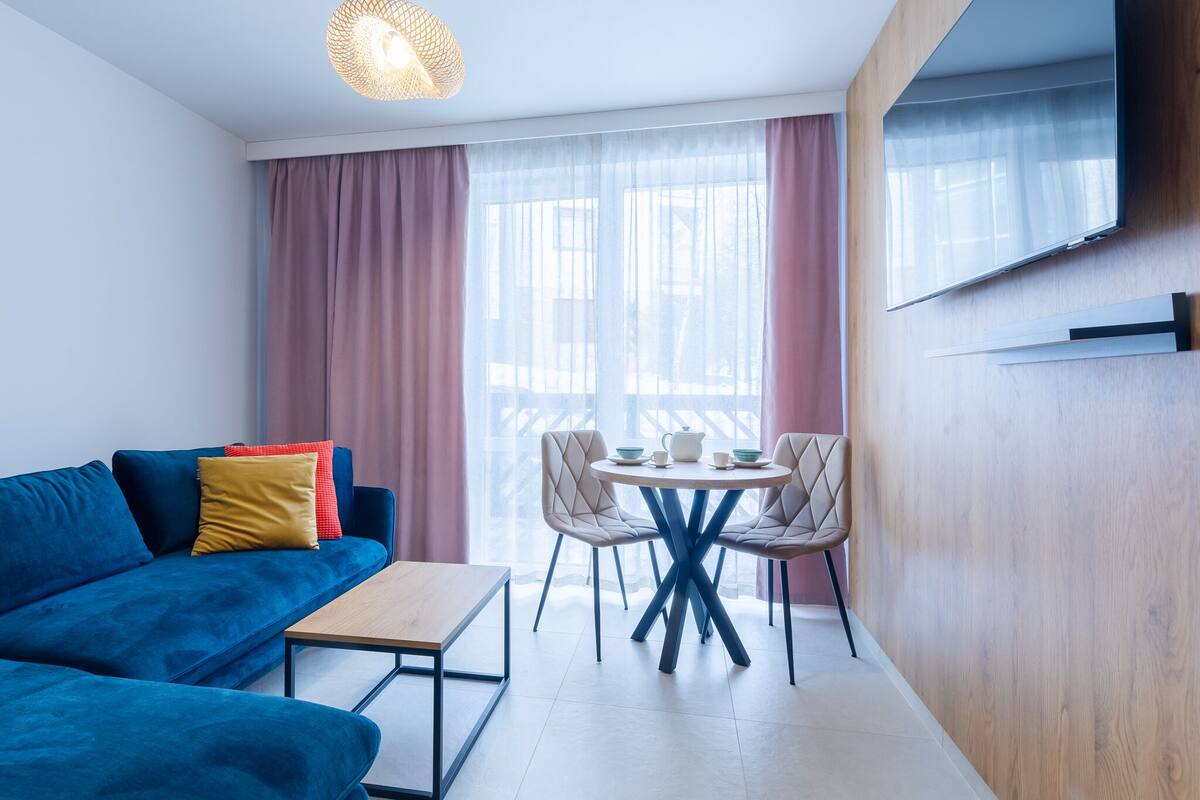 A modern room with a blue sofa, a small table, a round dining table with chairs, and a TV mounted on the wall.
