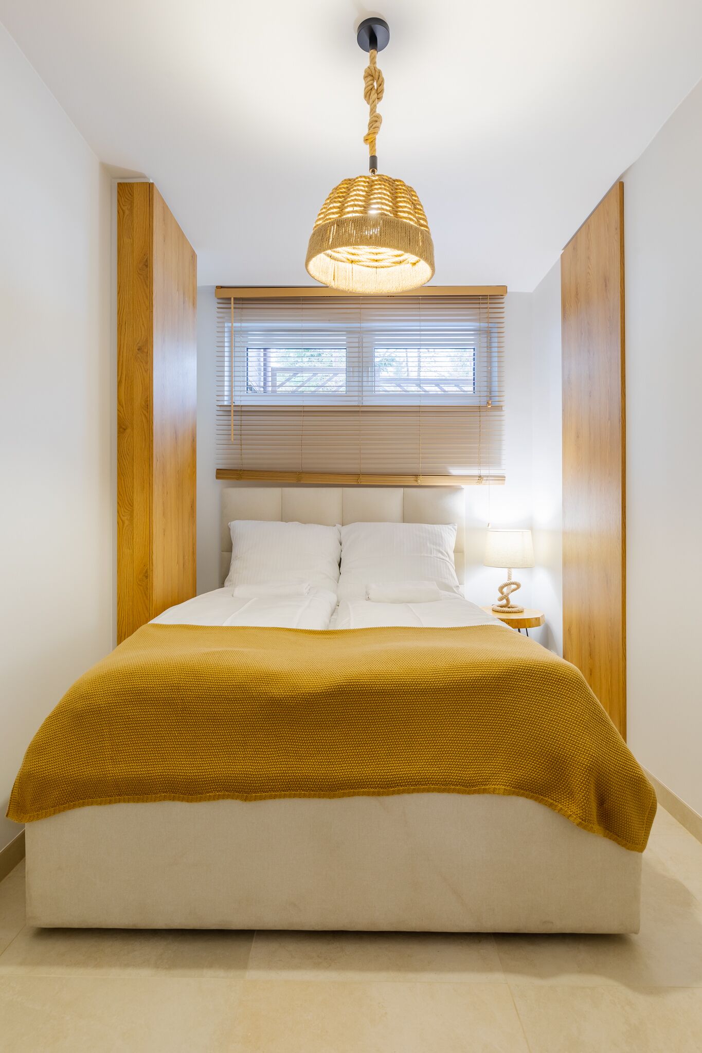Cama bem arrumada com manta cor mostarda, cabeceira de madeira e luminária pendente.