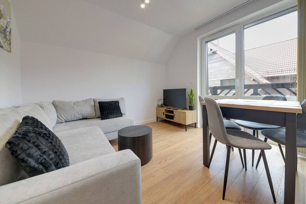 A modern living room with a sofa, a coffee table, a TV on a wooden stand, and a dining area with a table and chairs.