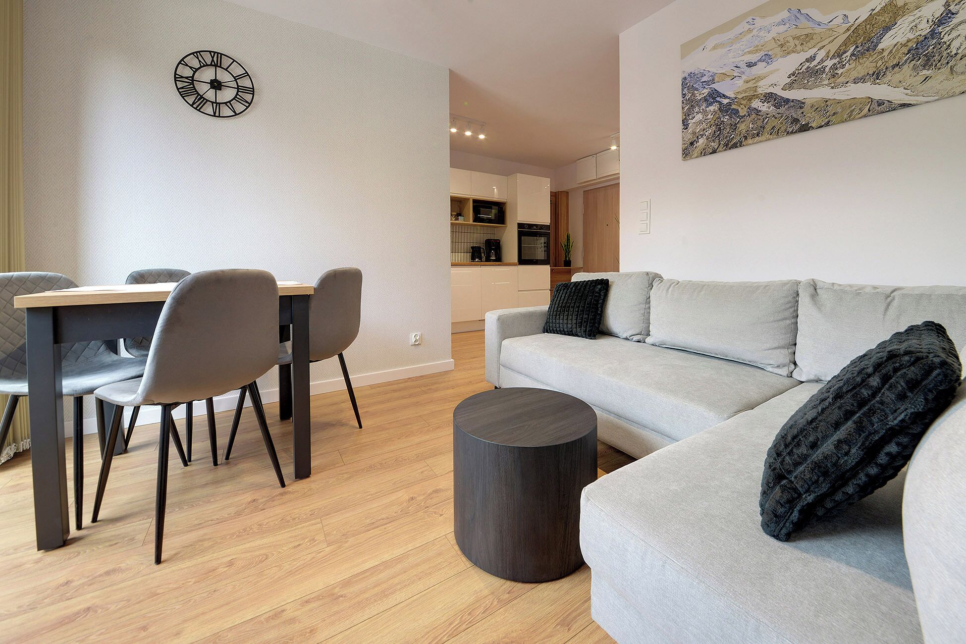 A modern living room with a grey sofa, a dining table with chairs, a round coffee table, and a kitchen in the background.