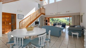 A spacious living area with a round dining table, blue cushioned chairs, a blue sofa, and a wooden staircase leading to an upper level.