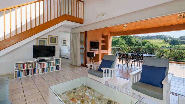 A spacious living area with a wooden staircase, a television, bookshelves, and outdoor seating with a view of greenery.