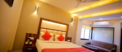 A hotel room with a large bed, a sofa, a ceiling fan, and a window with an air conditioner.