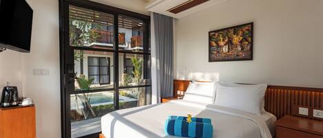 Ein Hotelzimmer mit einem großen Bett, einem Fernseher, einem Schreibtisch mit Kaffeemaschine und einem Balkon mit Blick auf ein Gebäude und Grünflächen.