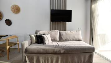 A modern living room with a sofa, a flat-screen TV mounted on the wall, and a dining table with chairs.