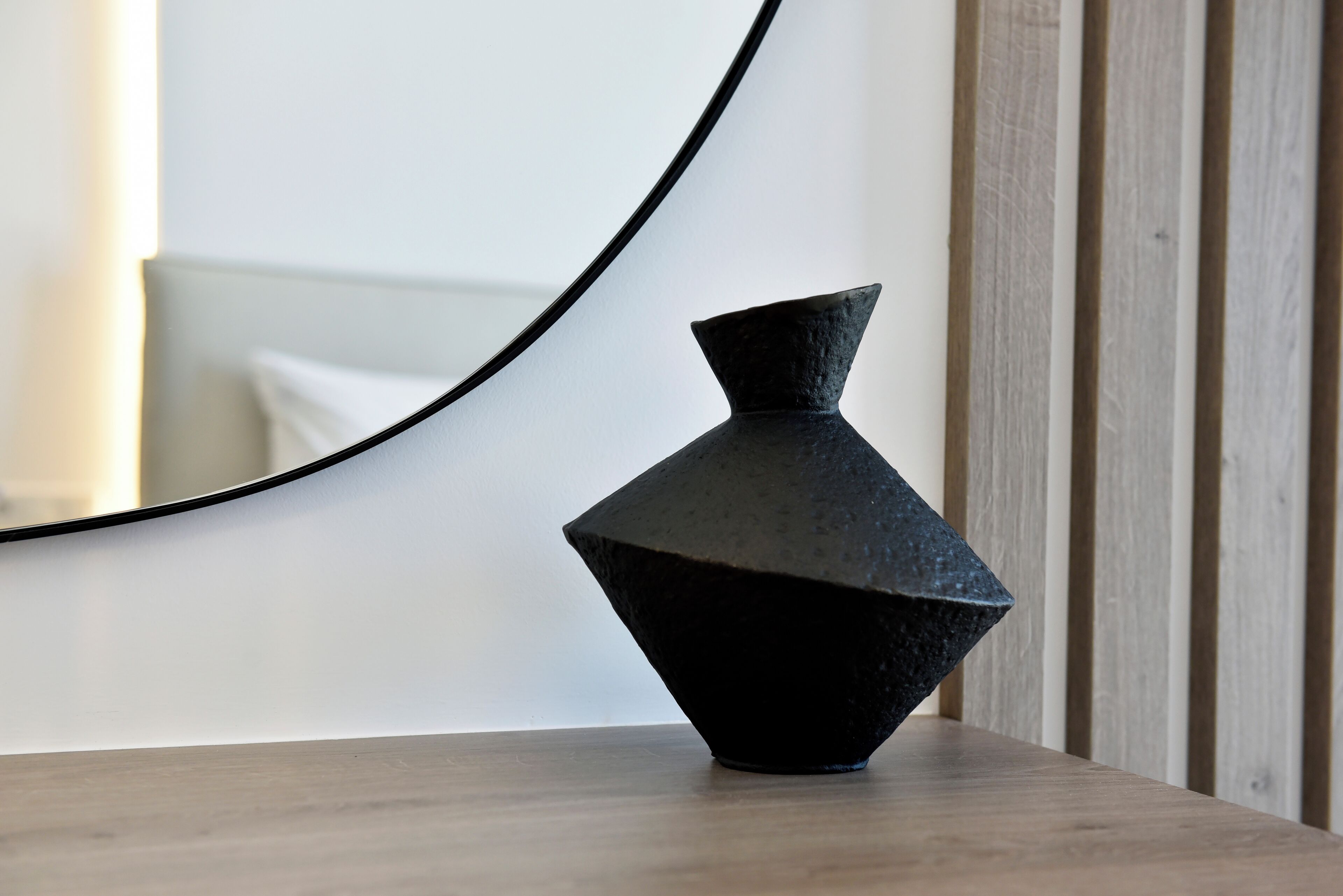 A black vase on a wooden table with a curved mirror reflecting a white wall and a wooden panel.