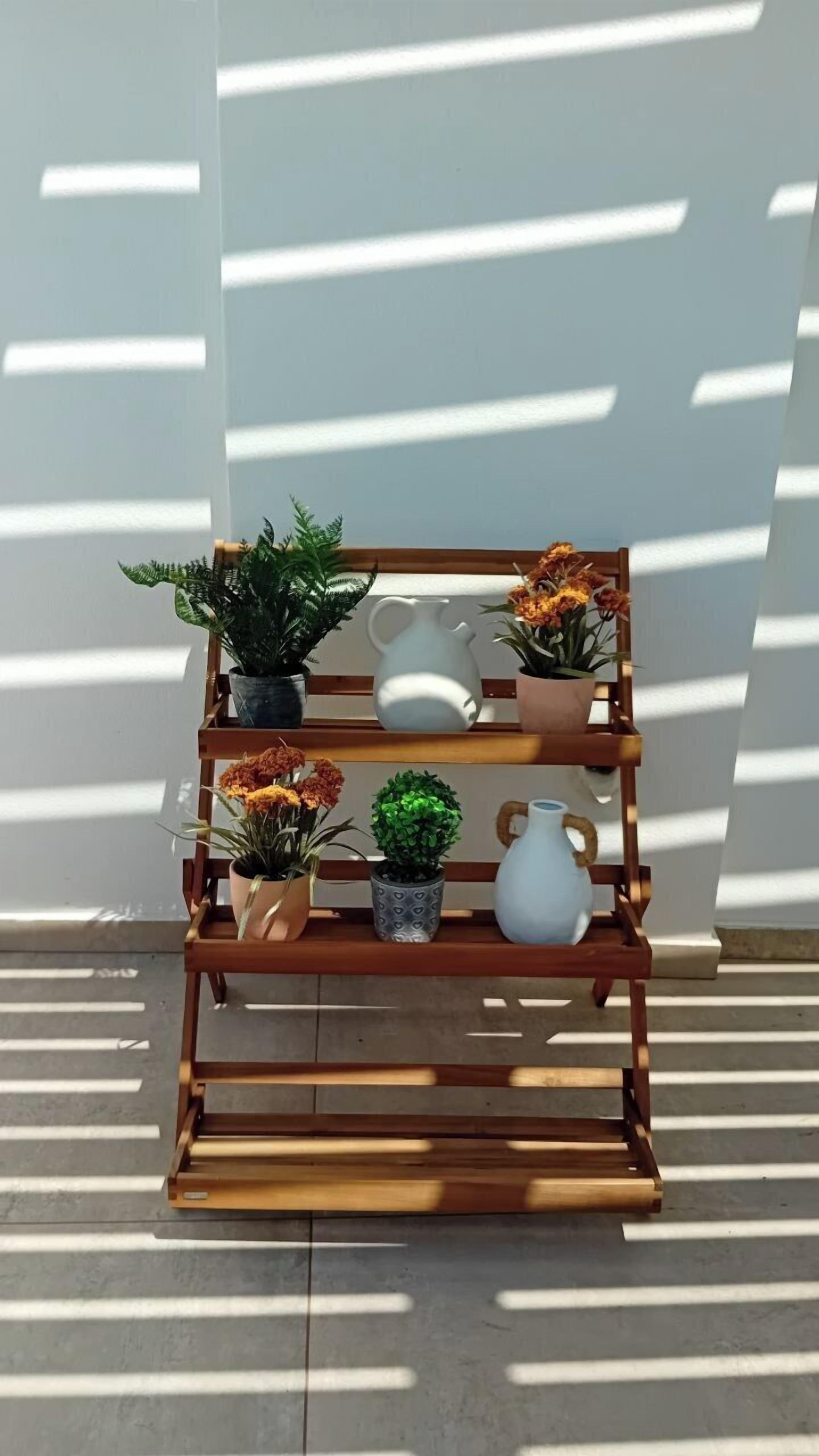 Un présentoir à plantes en bois, orné de différentes plantes en pot et d’objets décoratifs, est placé contre un mur où se projettent des ombres.