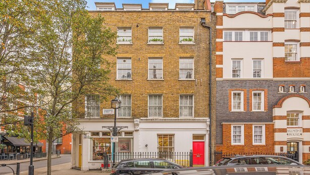 Une rangée d’immeubles à plusieurs étages, dont les façades sont faites d’un mélange de briques et de crépi, avec une porte rouge et une devanture visible, ornée d’une enseigne.