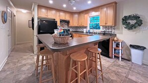 A kitchen with wooden cabinets, a black refrigerator, a large island with stools, and a window with a view. - Colston Creek- Nestled in the Trees with a beautiful creekside space (Bryson City)