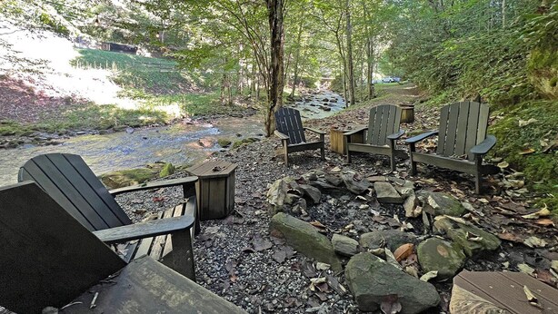 A serene outdoor seating area with Adirondack chairs and a small table, surrounded by rocks and trees, overlooking a stream. - Colston Creek- Nestled in the Trees with a beautiful creekside space (Bryson City)