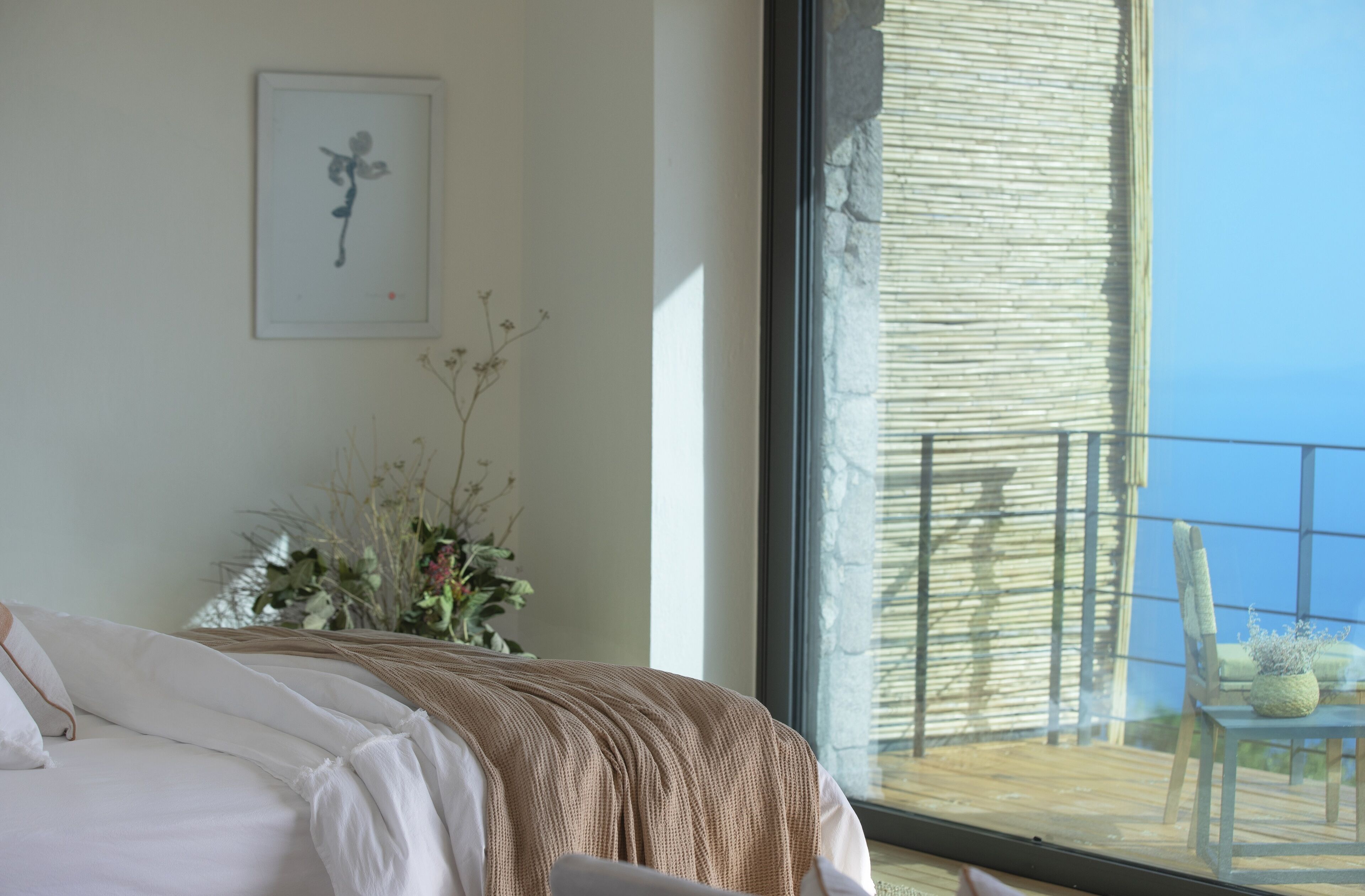 Een slaapkamer met een groot raam, een bed met witte lakens, een kunstwerk in een lijst aan de muur en een balkon met een stoel en een tafel.
