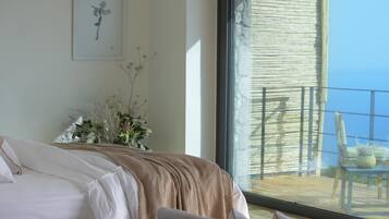 Een slaapkamer met een groot raam, een bed met witte lakens, een kunstwerk in een lijst aan de muur en een balkon met een stoel en een tafel.