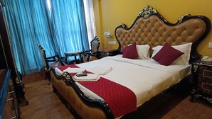A bed with a red quilt, a remote control, and a book on it. There is a nightstand with a lamp and a phone. The room has wooden floors and blue curtains. - N k Grand Days hotel (Port Blair)