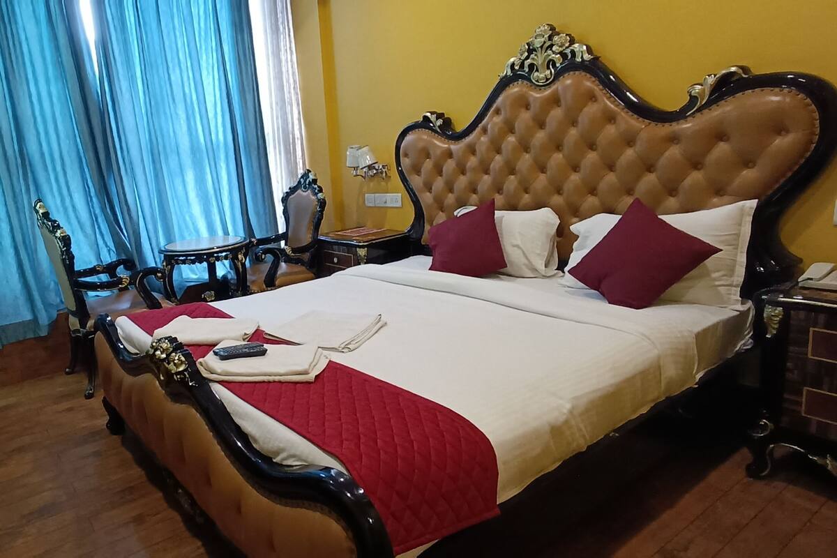 A bed with a red quilt, a remote control, and a book on it. There is a nightstand with a lamp and a phone. The room has wooden floors and blue curtains.