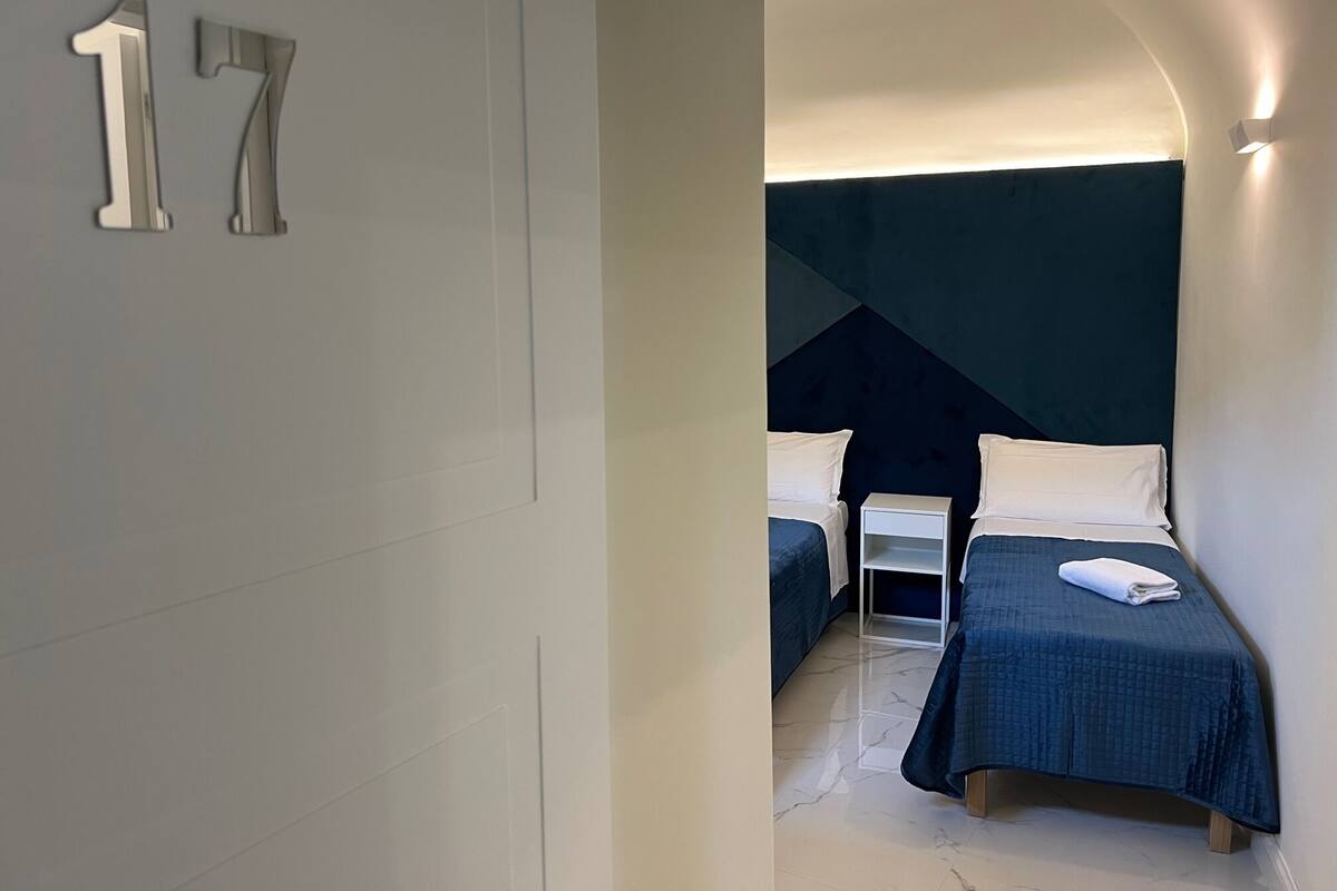 A modern hotel room with a large bed, a smaller bed, and a nightstand. The room has a minimalist design with a large number '17' on the wall.