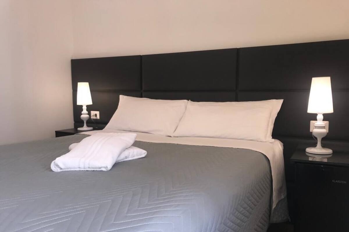 A neatly made bed with a grey quilt, two white pillows, and a folded white towel on the bed. There are two bedside tables with white lamps.