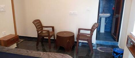 A small, compact room with a bed, two chairs, a small round table, a sink, and a blue trash bin.