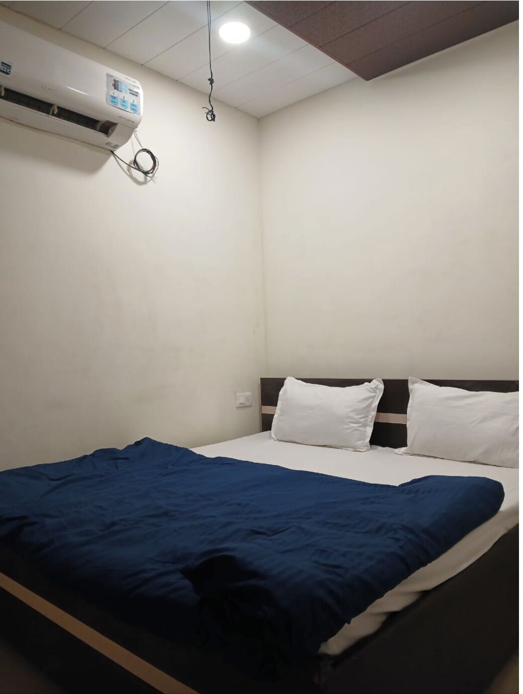 A hotel room with a bed, white walls, a ceiling-mounted air conditioner, and a visible power outlet.