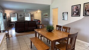 A dining area with a wooden table and chairs, a bench, and a potted plant. There is a sofa and a chair in the background.