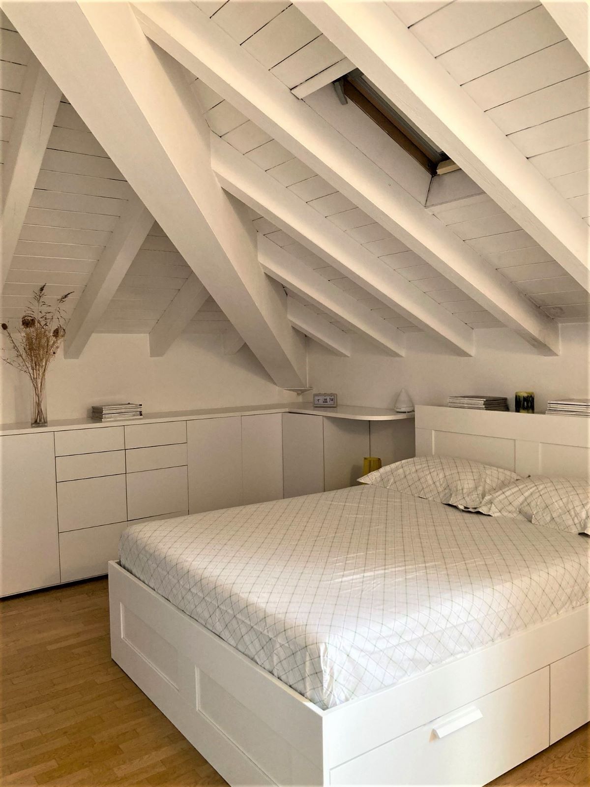 A bedroom with a sloped ceiling, white furniture, a bed with a checkered bedspread, and a small bedside table with a lamp.