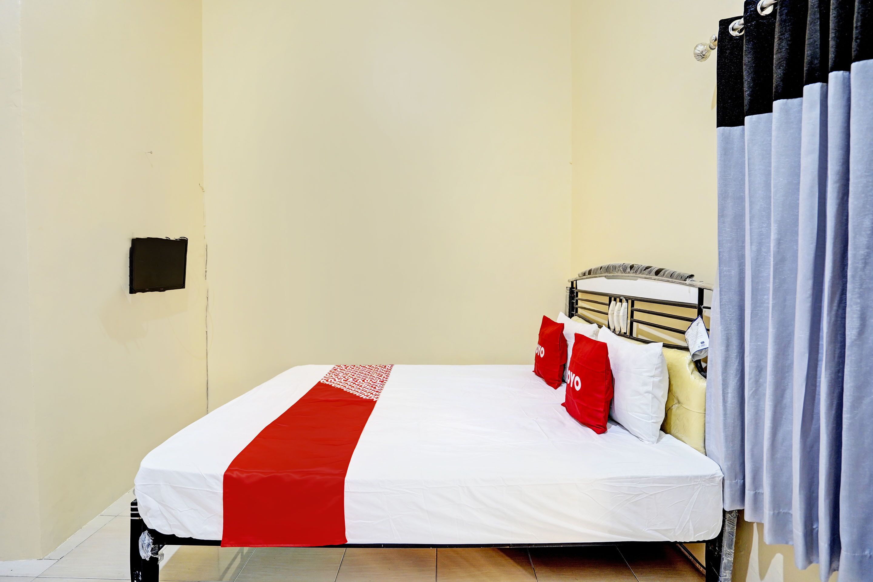 Una cama individual con colcha blanca y roja, cabecera y ventana con cortinas.