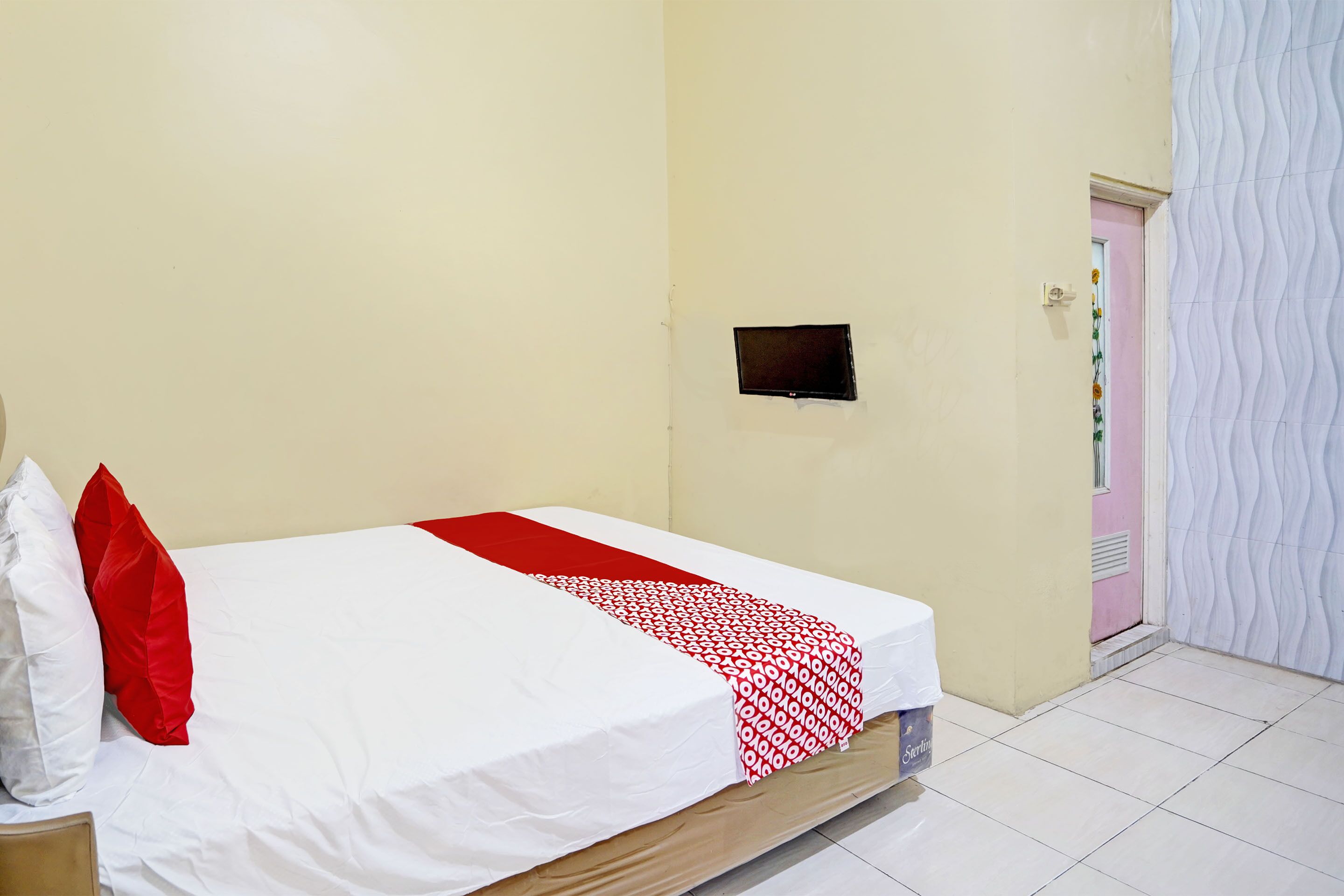 Una cama bien hecha con ropa de cama blanca y roja, un televisor negro pequeño montado en la pared y un piso de baldosas.