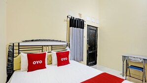 Un lit bien fait, recouvert d’une couverture blanche et rouge, avec deux oreillers ornés du mot « OYO », une tête de lit noire et un petit bureau avec une chaise placée à droite.