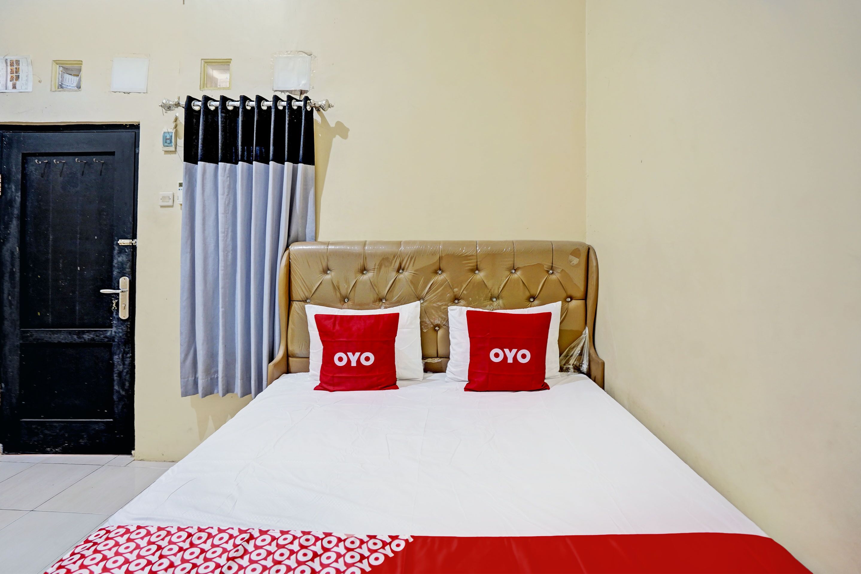 A hotel room with a bed, two red pillows with the word "OYO" on them, a black door, and a curtain.