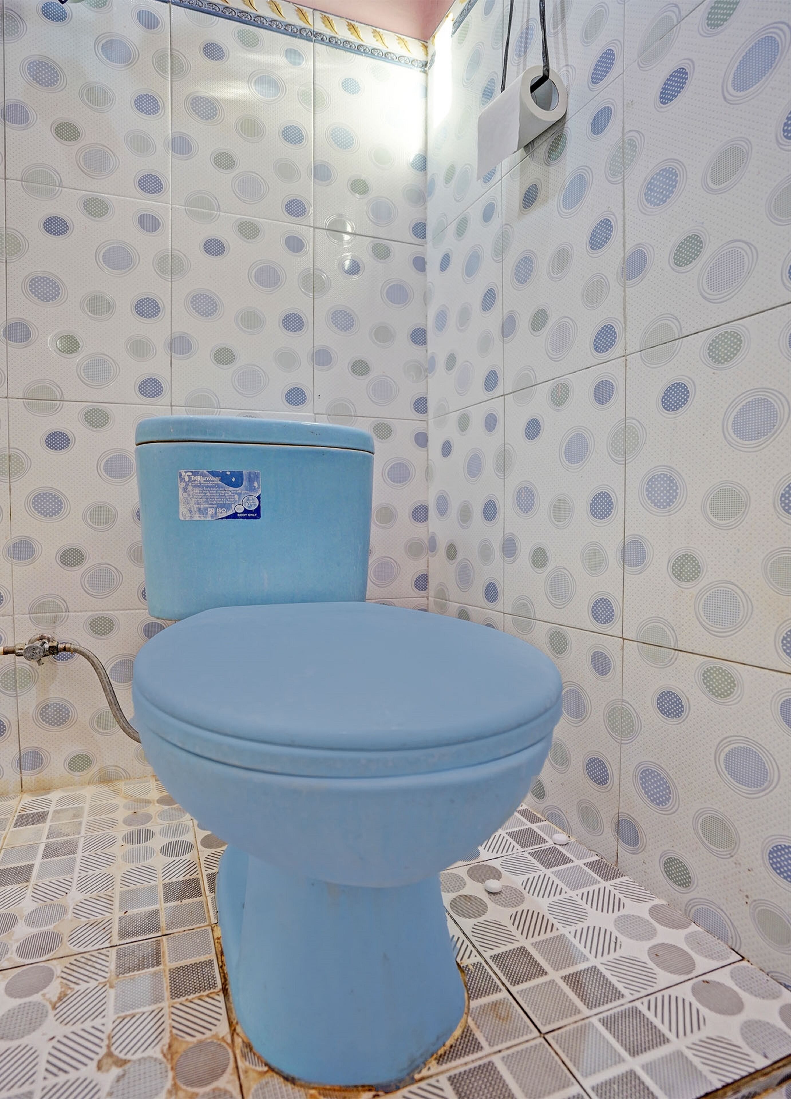 A bathroom with a blue toilet, patterned tiles, and a visible water pipe.