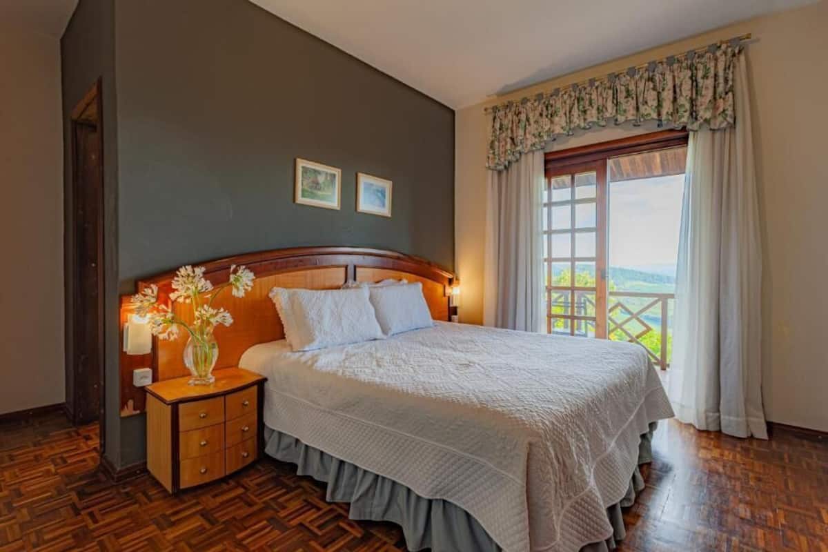 Un dormitorio con cabecera de madera, una cama grande con ropa de cama blanca, una mesita de noche con un florero y una ventana con vistas a zonas verdes.
