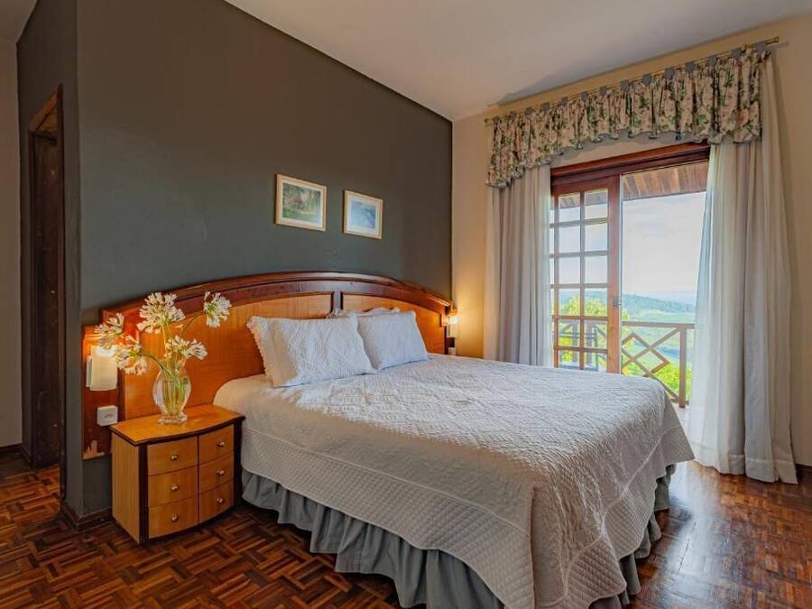 A bedroom with a wooden headboard, a large bed with white bedding, a nightstand with a vase of flowers, and a window with a view of greenery.