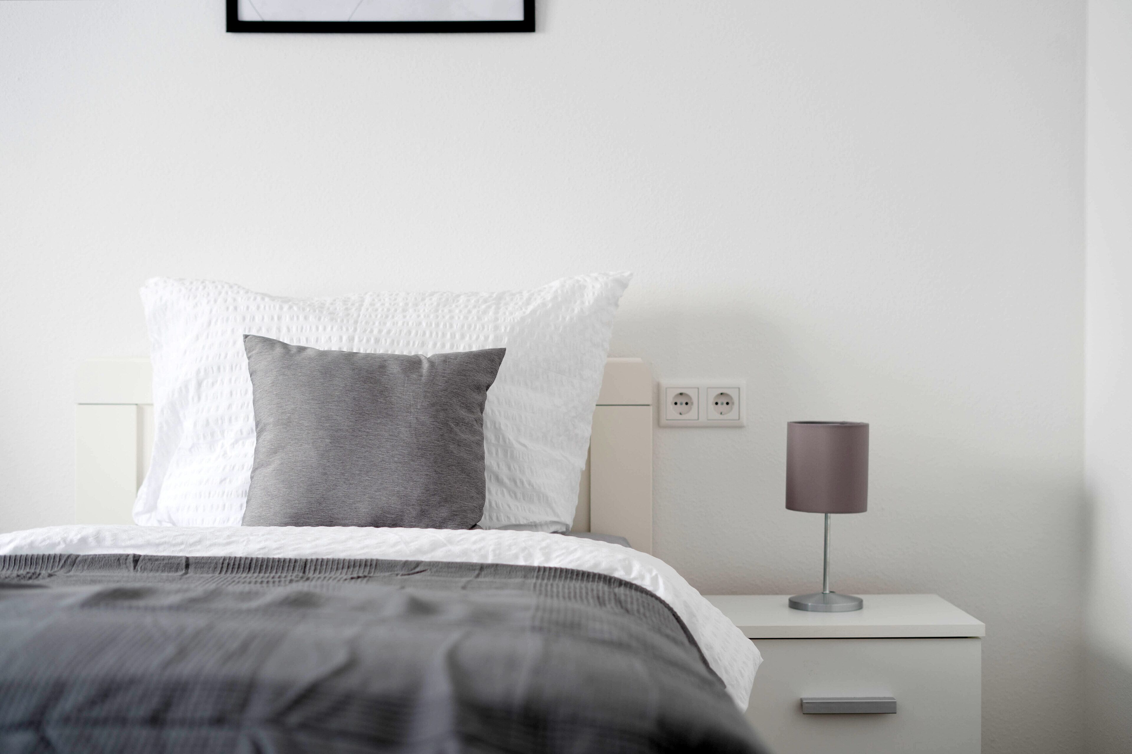 Ein ordentlich bezogenes Bett mit einer grau-weißen Tagesdecke, einer Nachttischlampe und einem weißen Nachttisch mit einer Schublade.
