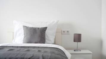 Ein ordentlich bezogenes Bett mit einer grau-weißen Tagesdecke, einer Nachttischlampe und einem weißen Nachttisch mit einer Schublade.