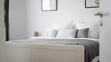 Een netjes opgemaakt bed met wit- en grijze beddengoed, een wit hoofdbord en een nachtkastje met een lamp.