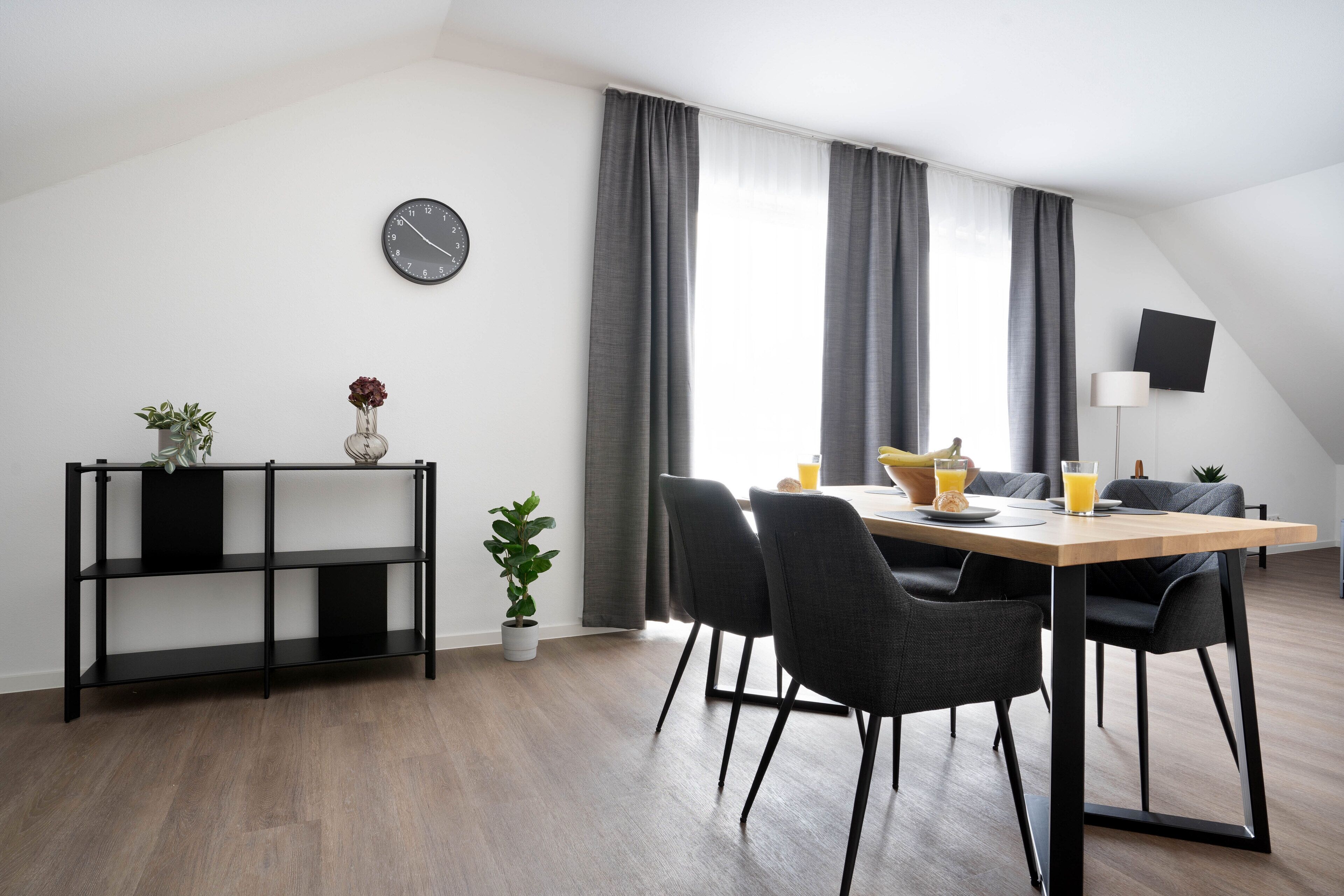 A modern dining area with a wooden table, four chairs, a black metal shelf with plants, a wall clock, and a TV mounted on the wall.