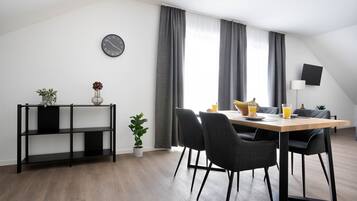 A modern dining area with a wooden table, four chairs, a black metal shelf with plants, a wall clock, and a TV mounted on the wall.