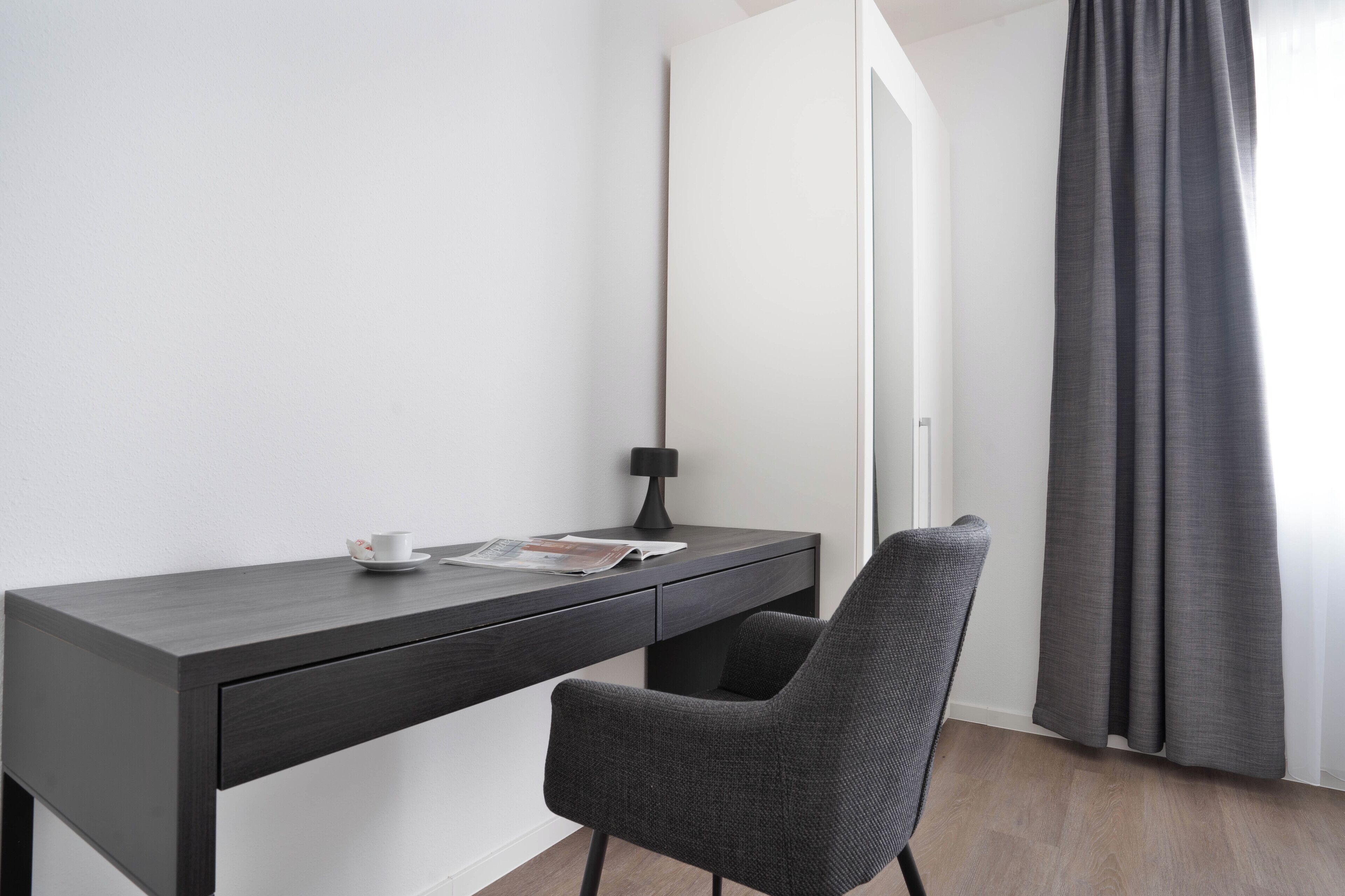 A modern room with a dark wooden desk, a chair, a lamp, and a window with curtains.