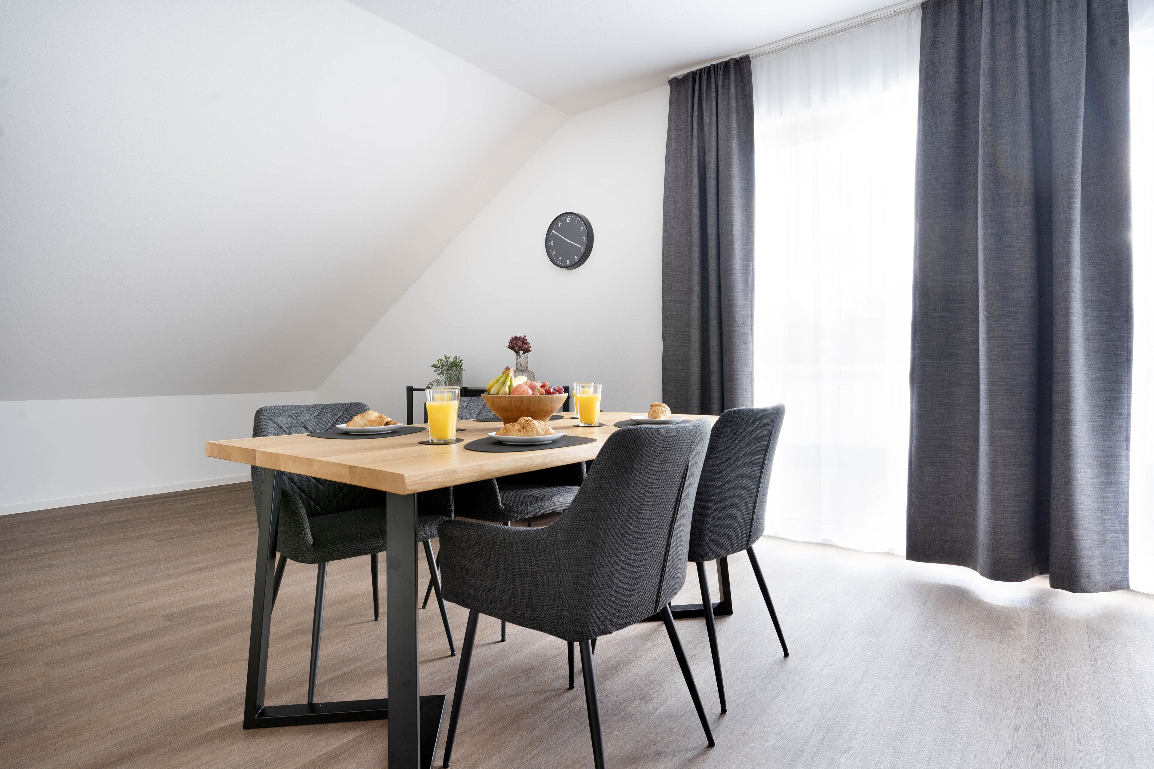 A modern dining area with a wooden table, four chairs, and a clock on the wall.
