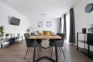 A modern dining area with a rectangular table, four chairs, a flat-screen TV mounted on the wall, and a coffee machine on a shelf. - Schöne Apartments in Lengerich (Lengerich)