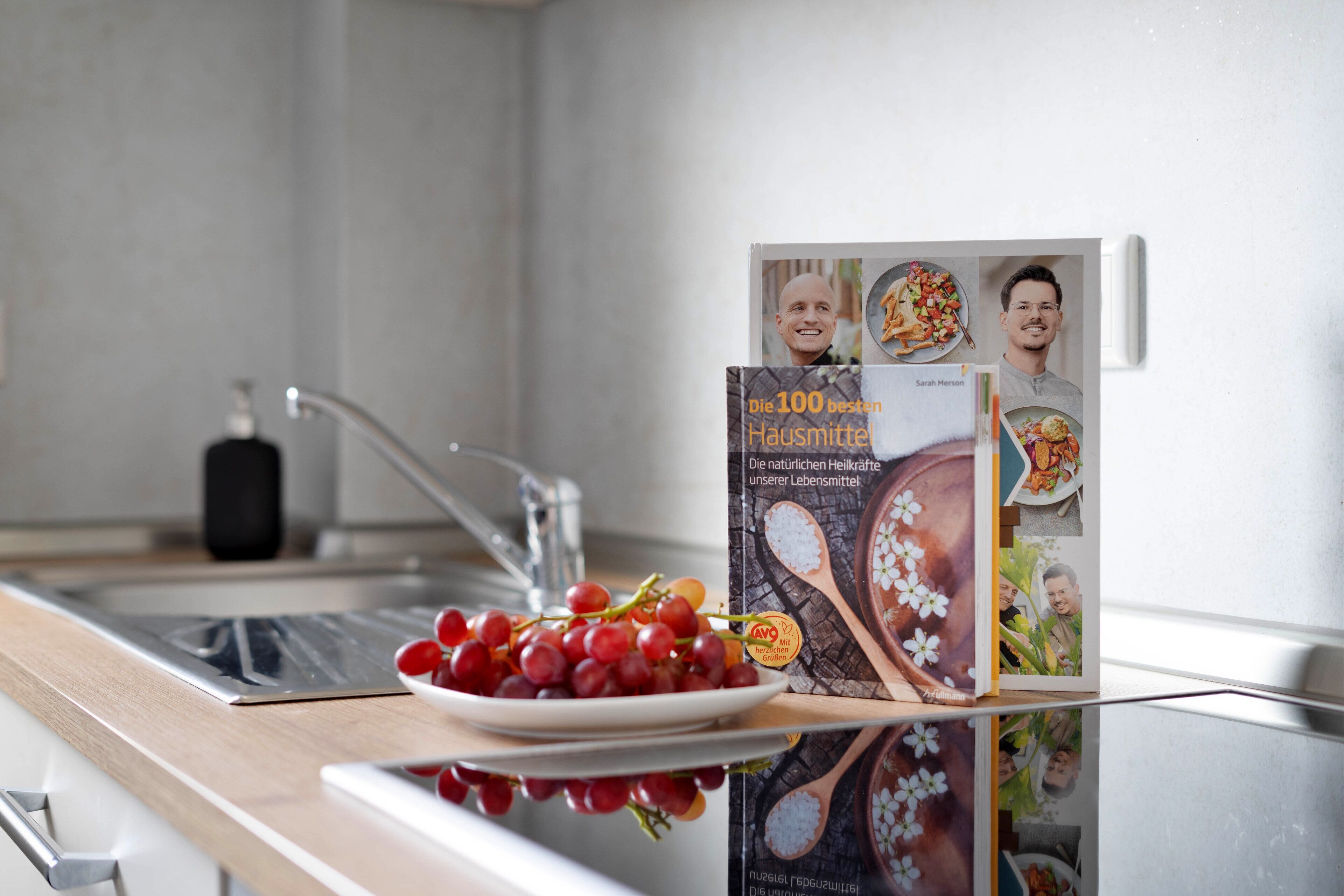 Un plan de travail de cuisine avec une assiette de raisins rouges, un robinet en acier inoxydable et un livre de cuisine intitulé « Les 100 meilleurs produits ménagers » sur une étagère.