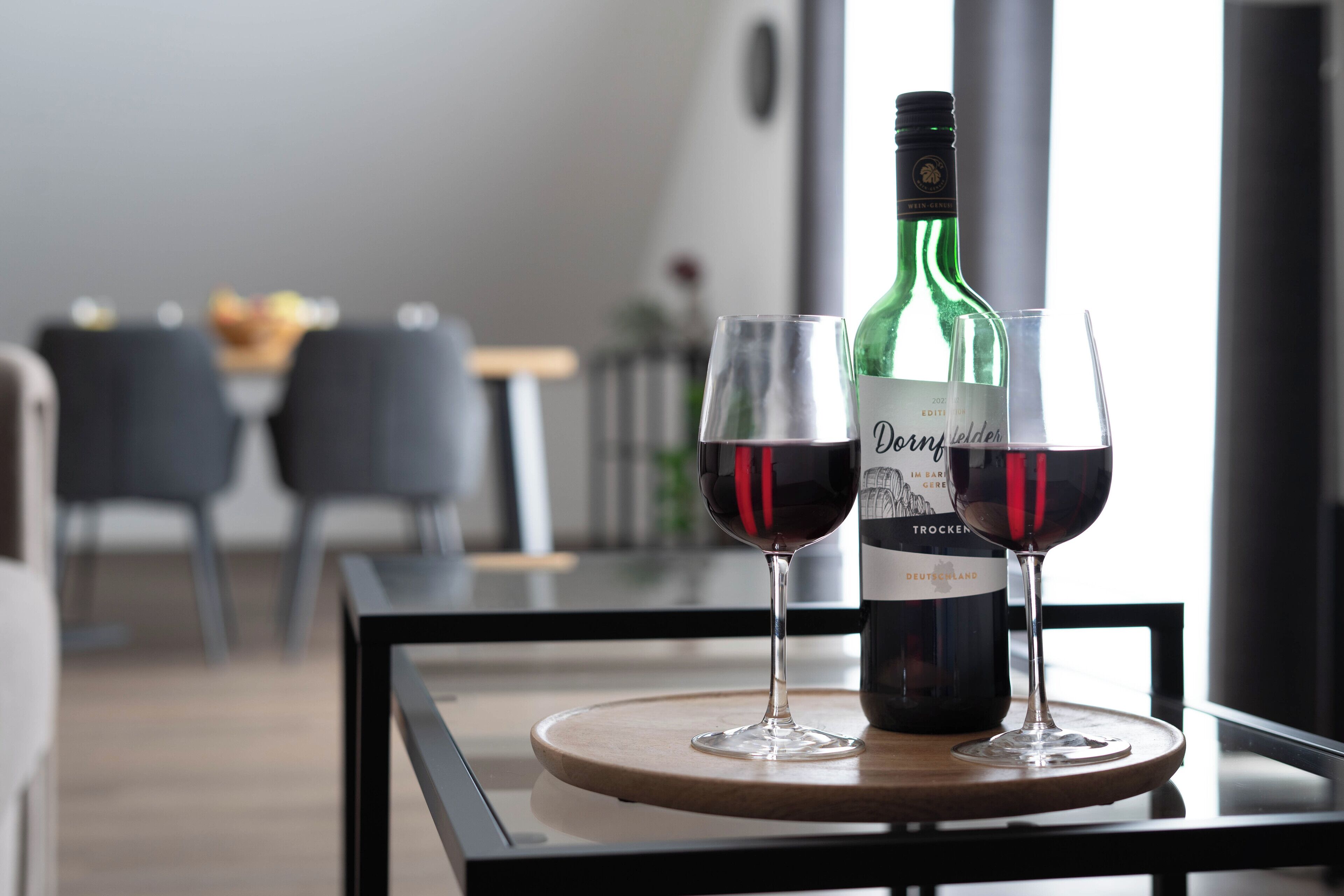 Una botella de vino Dornfelder y dos copas sobre una bandeja en un comedor moderno, con vista a una mesa y sillas al fondo.