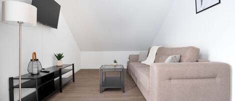 Een moderne woonkamer met een bank, een aan de muur bevestigde televisie, een vloerlamp en een klein bijzettafeltje met een plantje.